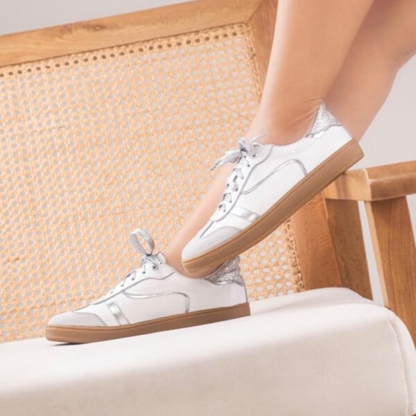 Tenis casuales de cuero blancos Alana con detalles plateados y suela color beige, sobre una silla de mimbre.
