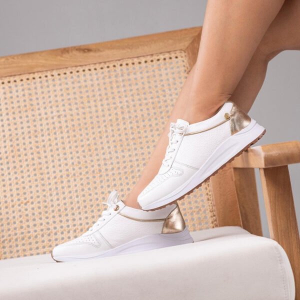 Tenis casuales de cuero blancos (modelo Snow) con paneles perforados, detalles metálicos dorados en el talón, también en una línea de este color en la parte alta, y suela gruesa blanca.