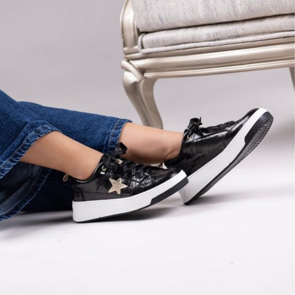 Tenis casuales de cuero negro con acabado brillante (modelo Estrella), cierre de cordones negros, una estrella decorativa dorada en el lateral y suela blanca con una delgada línea negra inferior.