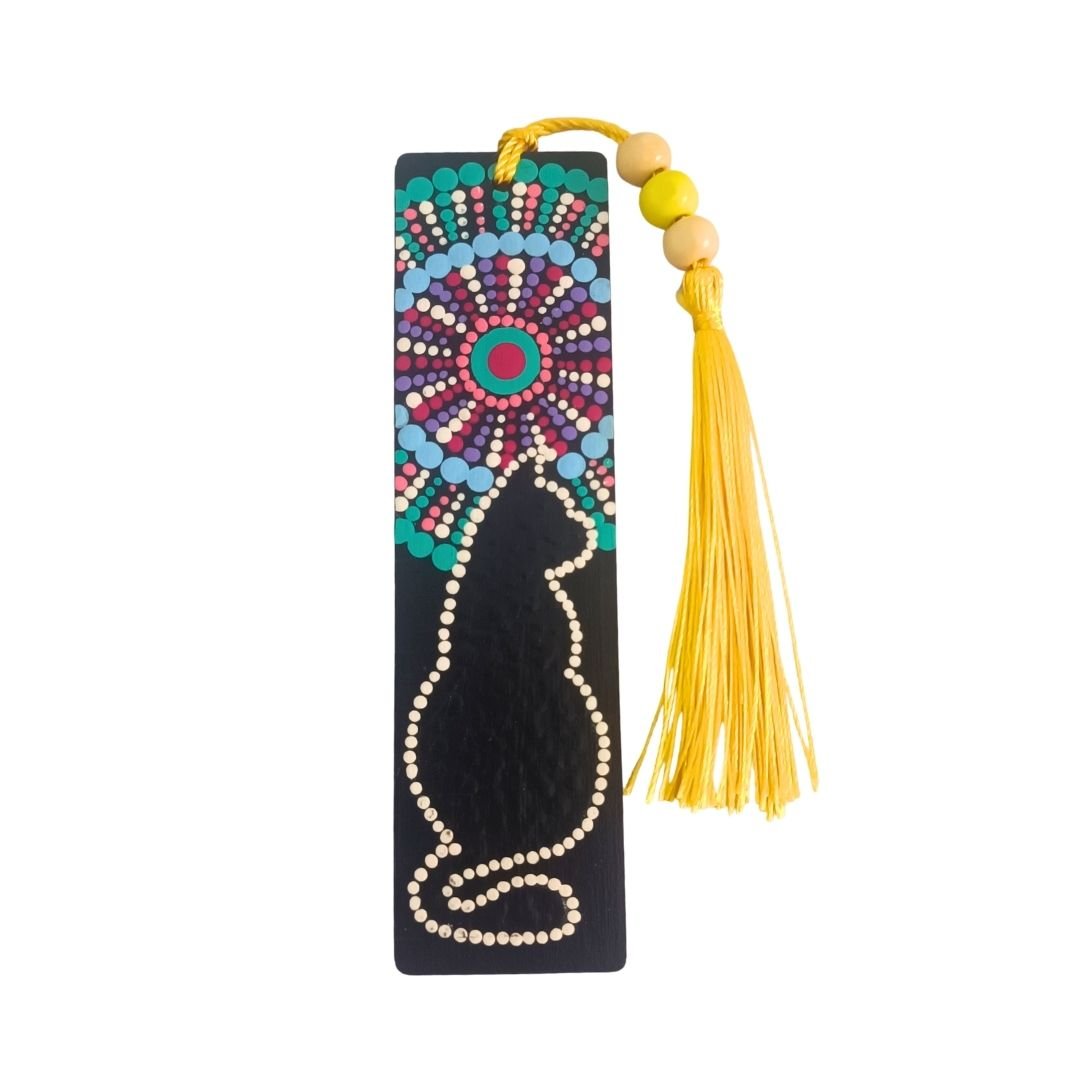 Separador de libros de madera negra con diseño de gato y mandala colorido, y una borla de hilo amarillo vibrante atada con cuentas de madera.