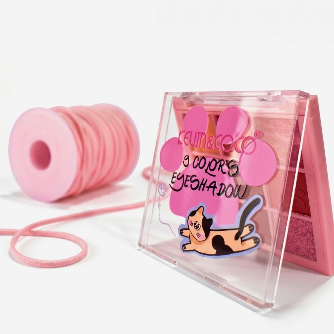 Composición de producto sobre fondo blanco. En primer plano y de perfil, una paleta de sombras de ojos de 9 colores con tapa transparente que lleva impresa una huella de gato fucsia, las palabras "9 COLORS EYESHADOW" y un dibujo de un gato tricolor jugando. Detrás, en el fondo, reposa un carrete redondo de hilo o cordón elástico de color rosa pastel.