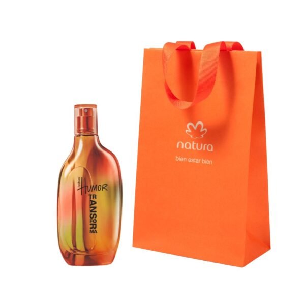 Un frasco de perfume Natura Humor Transforma de vidrio con tonalidades degradadas en naranja, amarillo y rojo, junto a una bolsa de regalo de papel color naranja brillante con asas de tela del mismo tono y el logotipo de Natura impreso en blanco.